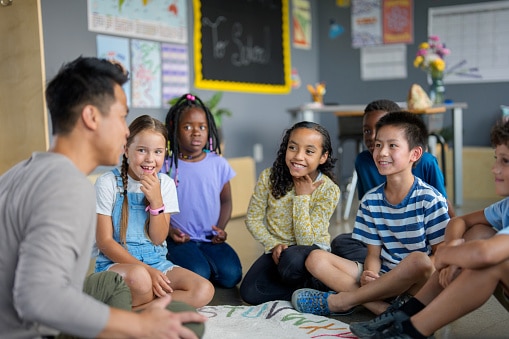Teach sitting on the floor with his students, telling a story.